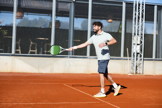 Male tennis player practicing a forehand on a sunlit clay court, extending his racket during an outdoor training session