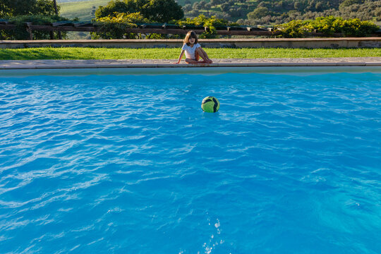 Young girl sitting by swimming pool edge with floating ball in foreground