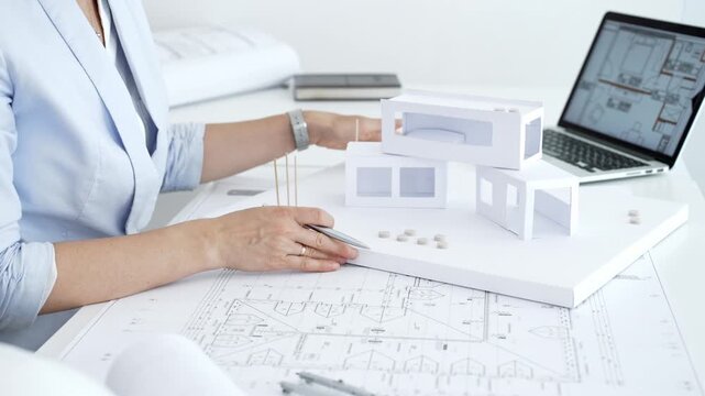 Architect actively building a paper architectural model on a desk strewn with blueprints and a laptop showing digital floor plans, capturing hands on design process