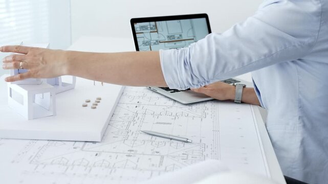 Architect actively building a paper architectural model on a desk strewn with blueprints and a laptop showing digital floor plans, capturing hands on design process