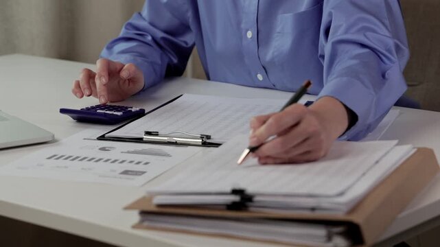 Person checking paperwork and documents at desk, writing notes beside calculator, clipboard, folders, and financial forms in an office setting. Hands of a woman doing payroll or calculating annual tax