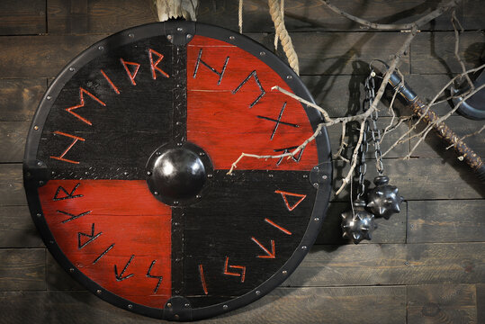 A red and black Viking style round shield decorated with runes hangs beside a flail with two spiked balls on a rustic wooden wall with bare branches.
