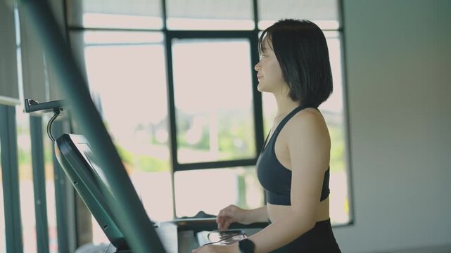 Young woman in black sports bra exercising on treadmill indoors near large window with natural light
