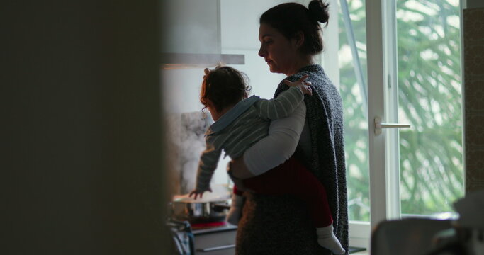 Mother carries toddler while cooking at stove in home kitchen, balancing meal preparation, childcare, household labor, multitasking demands and devoted everyday parenting
