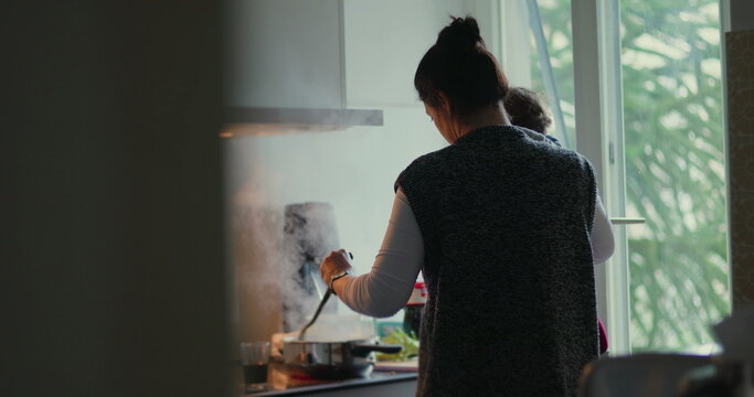 Busy parent stirs pot while holding young child on her hip in home kitchen, combining family cooking, childcare, mental load and modern caregiving responsibility