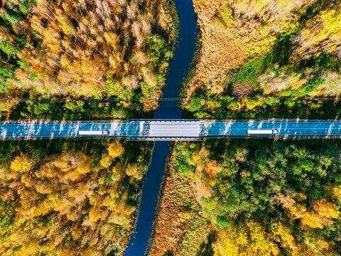 Aerial view of logistics trucks driving across river bridge on autumn forest highway route