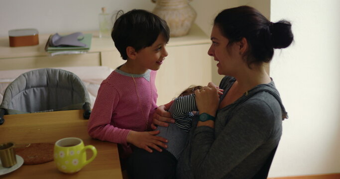 Breastfeeding mother talks with older son while nursing toddler at the breakfast table at home, capturing candid family interaction, maternal care and sibling closeness