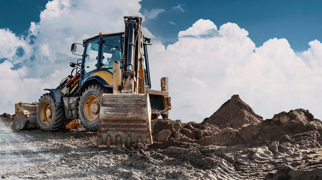 A backhoe loader uses its front bucket to move soil at a construction site, leaving tire tracks across the ground during daytime