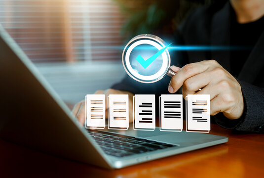 Business person holding a magnifying glass examining a quality checkmark icon over a row of digital documents on a laptop. Concept of compliance, audit, and quality control.