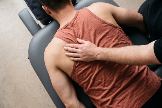 Overhead view of a male physiotherapist massaging and manipulating the shoulder and back of an athletic male patient lying face down on a medical massage table in a clinic