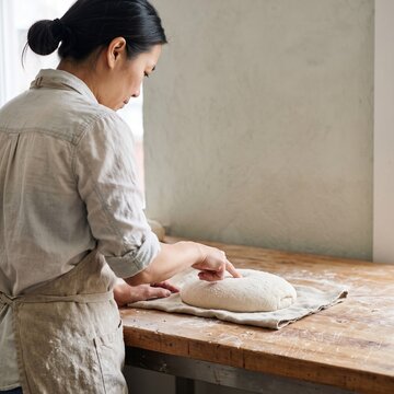 Asian female baker wearing a linen apron testing risen sourdough bread dough by poking it with her finger on a rustic wooden table