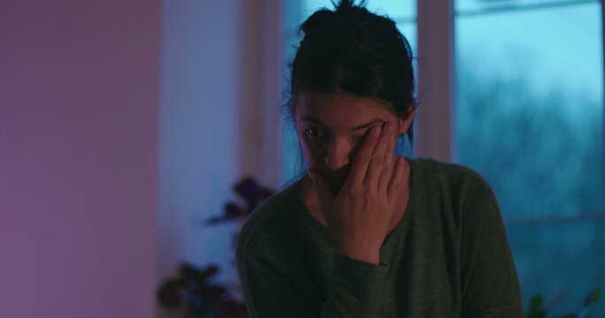 Exhausted woman rubs her eyes then rests her chin on her hand beside a window at home, showing fatigue, burnout and emotional strain from prolonged stress and overwork
