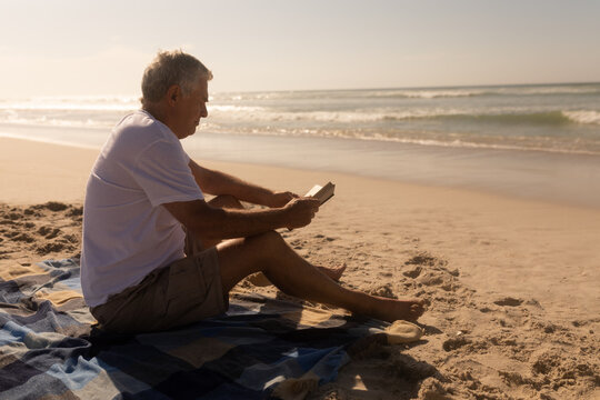 Senior man reading a book while relaxing on picnic blanket at beach