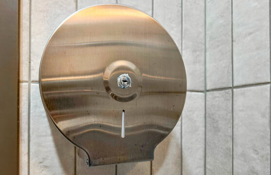 Wall mounted stainless steel toilet paper dispenser on tiled restroom wall. Public restroom hygiene and sanitation fixture. Commercial washroom supplies and facility maintenance