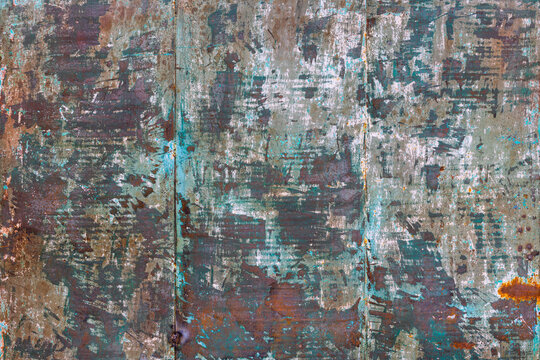 Close-up view of a weathered metal surface displaying extensive rust and peeling paint in teal, white, and dark brown. The rough texture features scratches and a vertical seam indicating joined panels