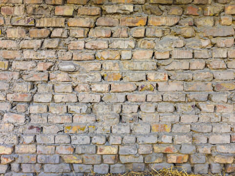 Detailed photograph of a weathered brick wall with irregularly shaped bricks in shades of tan, gray, and reddish-brown. Visible mortar and dry grass at the base enhance the texture.