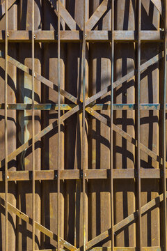 Close-up view of a rusty metal gate featuring vertical bars and diagonal crossbars. Strong sunlight creates geometric shadows on the corrugated metal background, enhancing texture and depth.