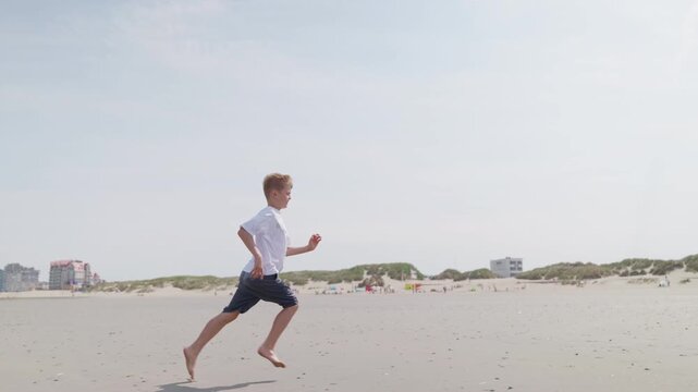 A boy running along wet beach sand dunes and buildings in background slow motion by the sea looks at the camera and laughs