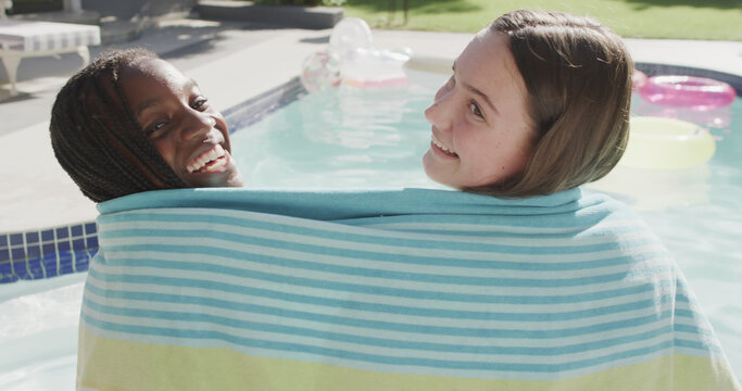 Two happy diverse teenage girl friends wrapped in towel sitting by sunny swimming pool