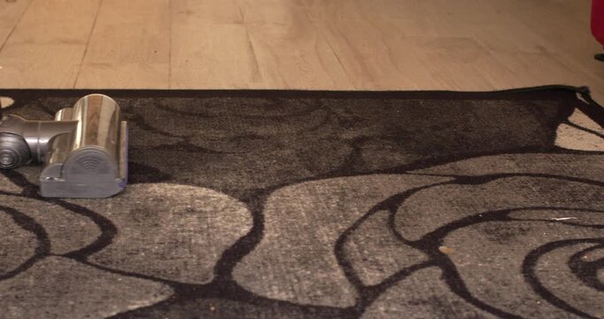 A vacuum cleaner on a dark gray rug with a black floral pattern, placed on a wooden parquet floor in a home interior. High angle shot.