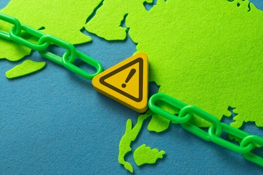 Yellow warning symbol and green chain arranged across a world map. Global supply chain disruption, regional logistics risk, trade route bottleneck, geopolitical hazard concept.