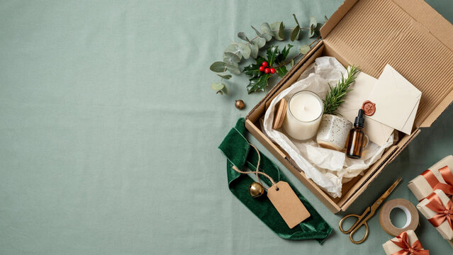 Premium Christmas Gift Packaging Flat Lay with Kraft Mailer Box, Candle, Ceramic Mug, Velvet Ribbon and Natural Holiday Greenery on Sage Green Background with Copy Space