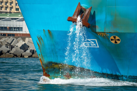 cargo ship bow anchor being raised with cascading water streaming down the turquoise hull at port with rocky breakwater behind