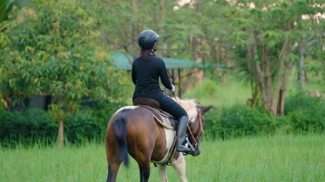 Woman wearing helmet riding a brown horse outdoors in nature with green trees and building background during daytime