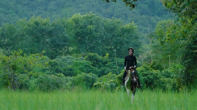 Asian Young woman riding horse through lush green meadow surrounded by tropical forest. Slow Motion.