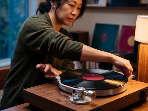 Middle aged Asian woman carefully placing a vinyl record onto a wooden turntable in a cozy living room illuminated by warm lamp light during the evening