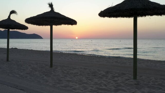 Thatched beach parasols silhouetted against sunrise over the sea in Mallorca, Spain