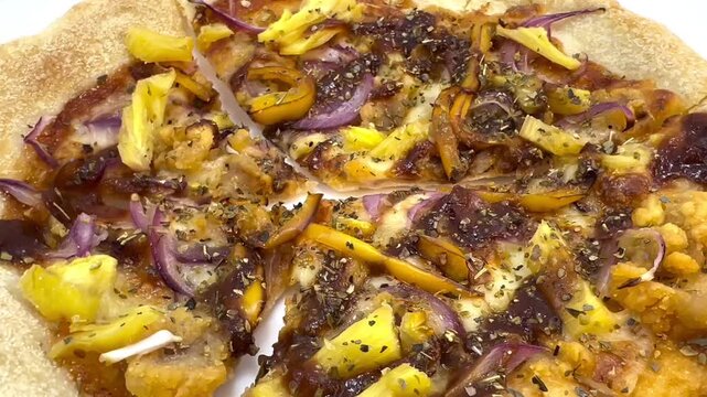 Top view of a round pizza cut into slices, topped with pineapple chunks, red onion, yellow bell pepper, cheese and dried herbs on a white surface.
