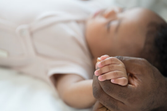 Close up of hands African American father gently holding tiny hands of newborn baby lying on white bed, baby care, paternal care, fatherhood, protection and healthcare bonding at home