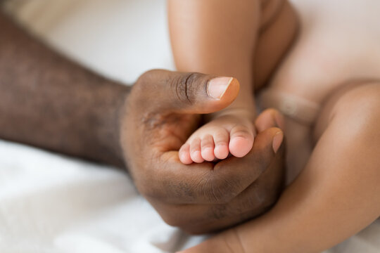 Hands of caring black father holding little feet of mixed face newborn infant on white bed, warmth, innocence, gentle touch, parenthood, baby care, healthcare bonding at home