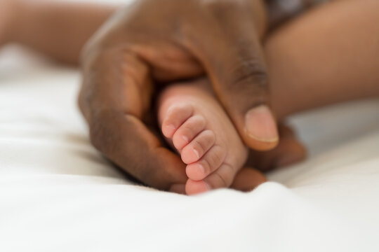 Hands of caring black father holding little feet of mixed face newborn infant on white bed, warmth, innocence, gentle touch, parenthood, baby care, healthcare bonding at home