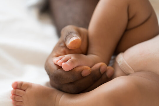 Hands of caring black father holding little feet of mixed face newborn infant on white bed, warmth, innocence, gentle touch, parenthood, baby care, healthcare bonding at home