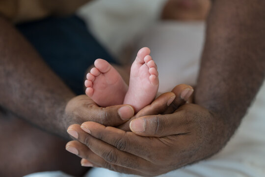 Close up of hands African American father gently holding tiny feet of newborn baby lying on white bed, tender moment of paternal care, fatherhood, protection, unconditional family love