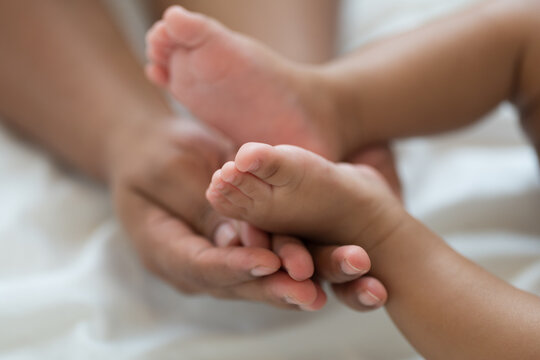 Close up of hands African American father gently holding tiny feet of newborn baby lying on white bed, tender moment of paternal care, fatherhood, protection, unconditional family love
