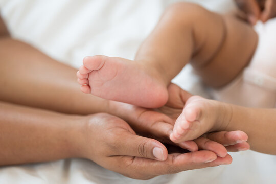 Close up of hands African American father gently holding tiny feet of newborn baby lying on white bed, tender moment of paternal care, fatherhood, protection, unconditional family love