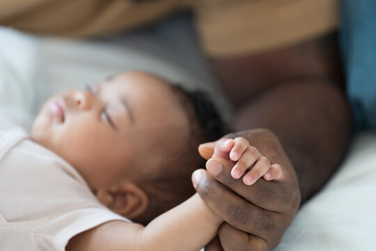 Close up of hands African American father gently holding tiny hands of newborn baby lying on white bed, baby care, paternal care, fatherhood, protection and healthcare bonding at home