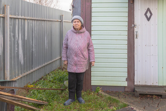 Elderly woman standing outside a modest house beside a metal gate. Senior woman at home in a residential yard. Aging, independent living and everyday life. Portrait of older adult