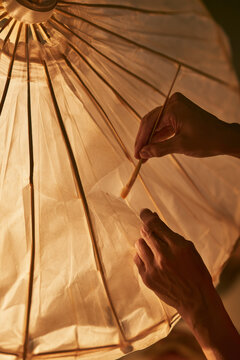 Artisan repairs a delicate tear in a paper parasol by hand in warm golden light