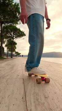 Vertical 9:16 detail footage of feet riding a skateboard along a waterfront promenade. Low ground-level perspective highlights motion, footwork, wheels and the relaxed coastal sunset atmosphere.