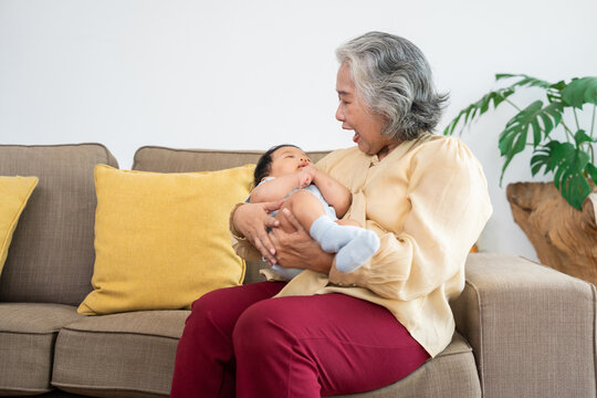 Happy Asian grandmother cradles her baby grandchild on a living room sofa, sharing a tender family moment in a bright home, ideal for parenting, childcare, and intergenerational love.