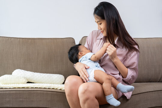 Asian mother sits on a sofa and looks lovingly at her baby resting across her lap, capturing an intimate at-home parenting moment for family, infant care, and maternity marketing