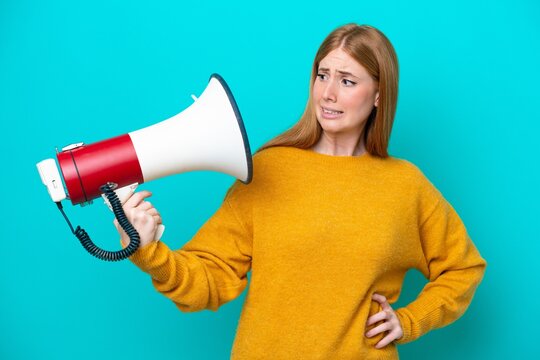 Young redhead woman isolated on blue background holding a megaphone with stressed expression