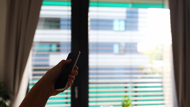 Hand holds a remote control to operate motorized window blinds in a modern apartment. Smart home automation allows convenient adjustment of daylight, privacy and indoor comfort
