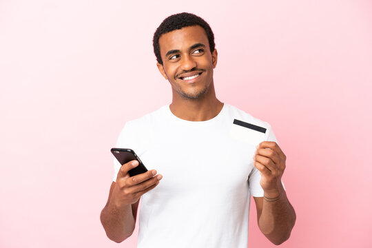 African American handsome man on isolated pink background buying with the mobile with a credit card while thinking