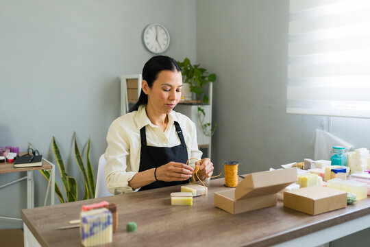 Woman creating artisan soap packaging, tying string around natural handmade bar for small business or craft shop