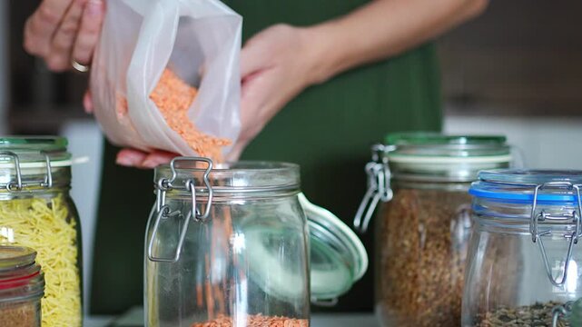 Woman pours red lentils from a plastic bag into a glass storage jar in the kitchen. Organizing dry food in reusable containers helps reduce packaging waste and keep the pantry tidy